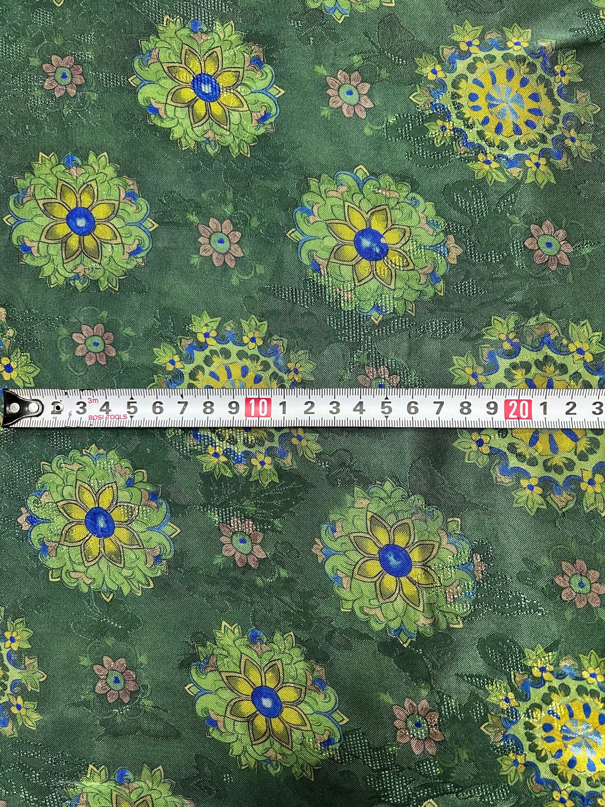 Close-up of dark green fabric with repeating yellow, lime green, and blue floral medallions, smaller dusty pink flowers, and a textured dotted background; a measuring tape crosses the center.