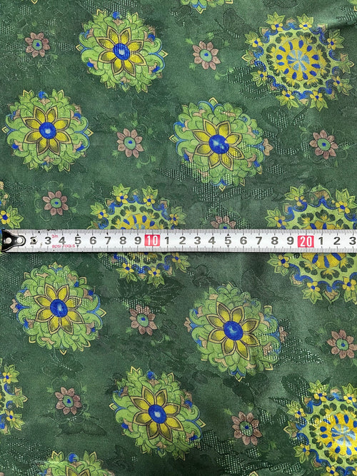 Close-up of dark green fabric with repeating yellow, lime green, and blue floral medallions, smaller dusty pink flowers, and a textured dotted background; a measuring tape crosses the center.