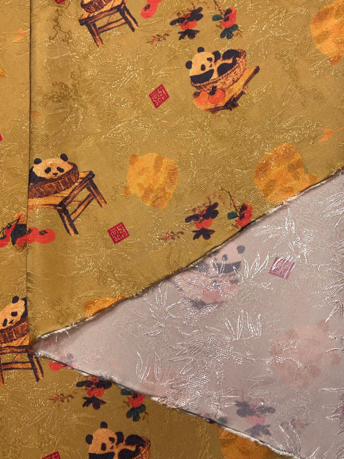Close-up of folded cloth with a mustard-gold side printed with pandas on chairs, red flowers and small red square motifs; the exposed reverse is dusty pink with white leafy designs.