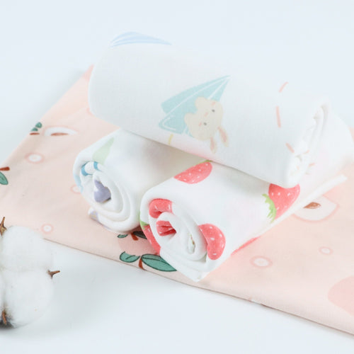 Close-up of three folded white cloths with pastel character and strawberry motifs, stacked on a pale pink printed cloth.