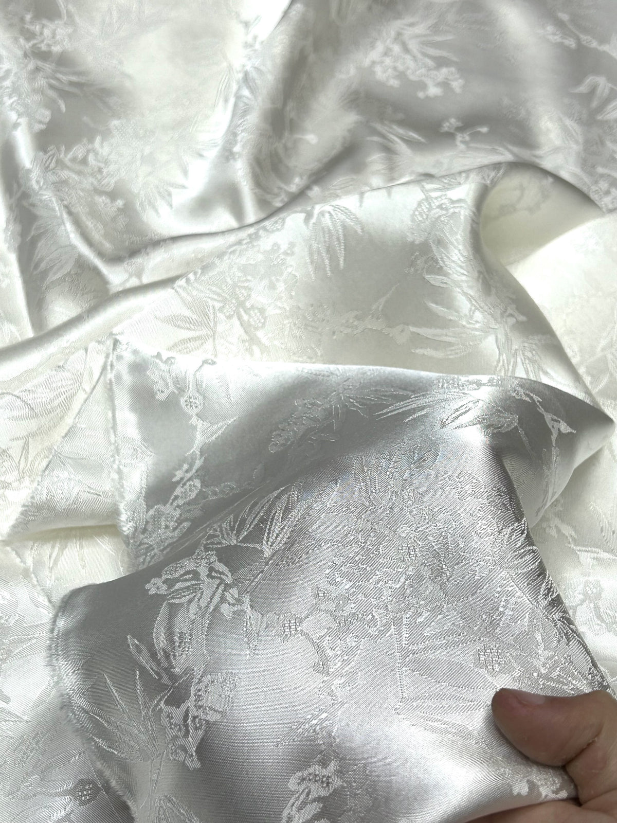Close-up of folded glossy ivory and silver cloth with an allover floral and leafy motif; a hand holds one corner.
