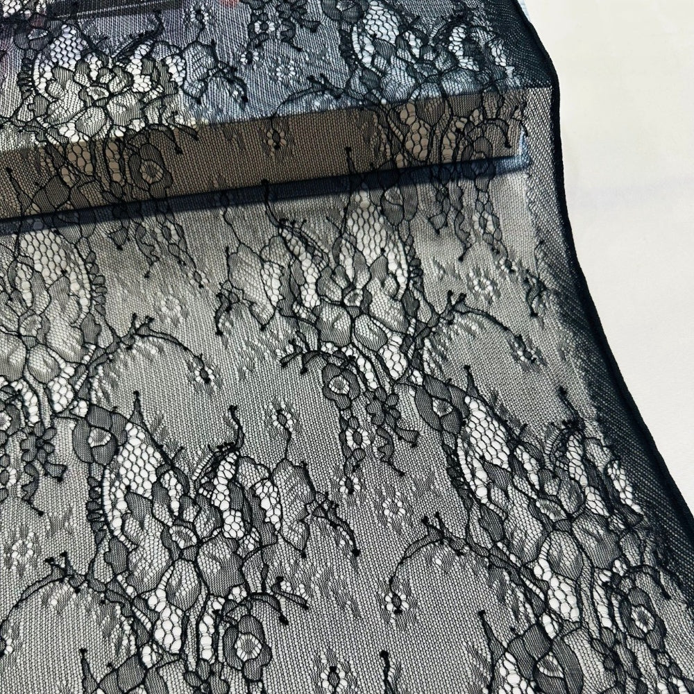 Black floral lace fabric with an open mesh ground and a narrow dark band, shown in a close view against a light background.