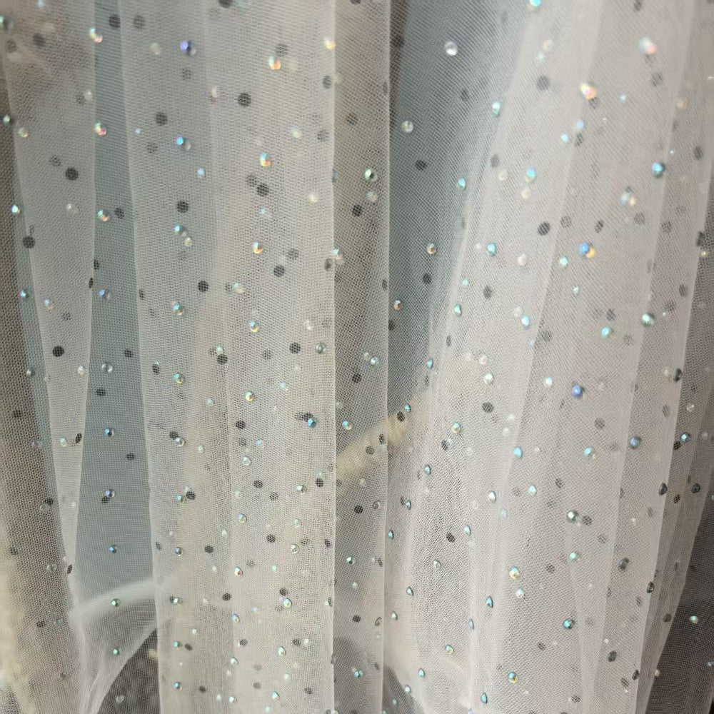 Ivory sheer mesh fabric hangs in loose vertical folds, dotted with small dark spots and scattered iridescent accents.