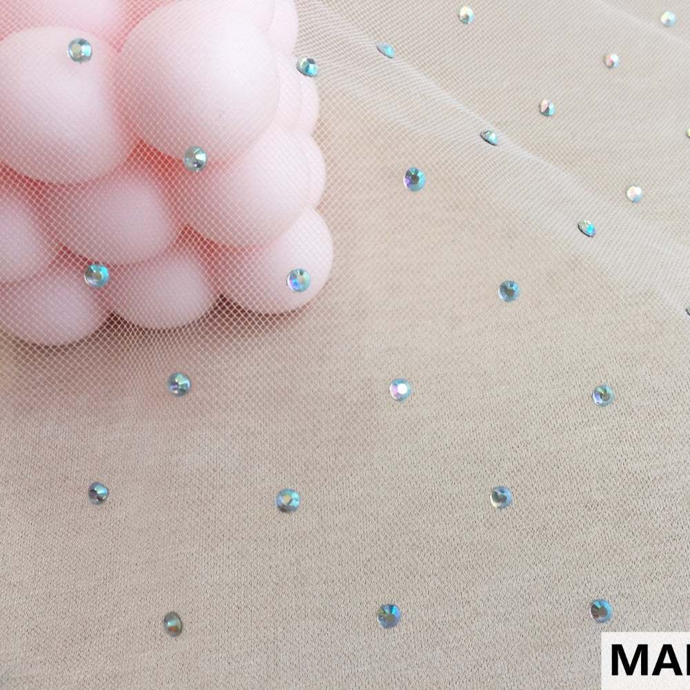 Close view of beige net fabric with small, spaced iridescent round embellishments. Pink rounded shapes are visible beneath the mesh at the upper left.