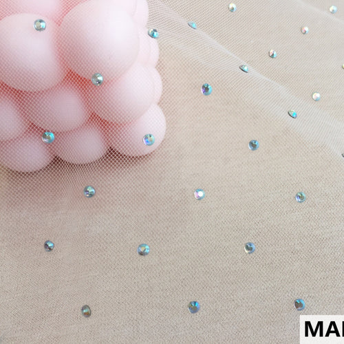 Close view of beige net fabric with small, spaced iridescent round embellishments. Pink rounded shapes are visible beneath the mesh at the upper left.