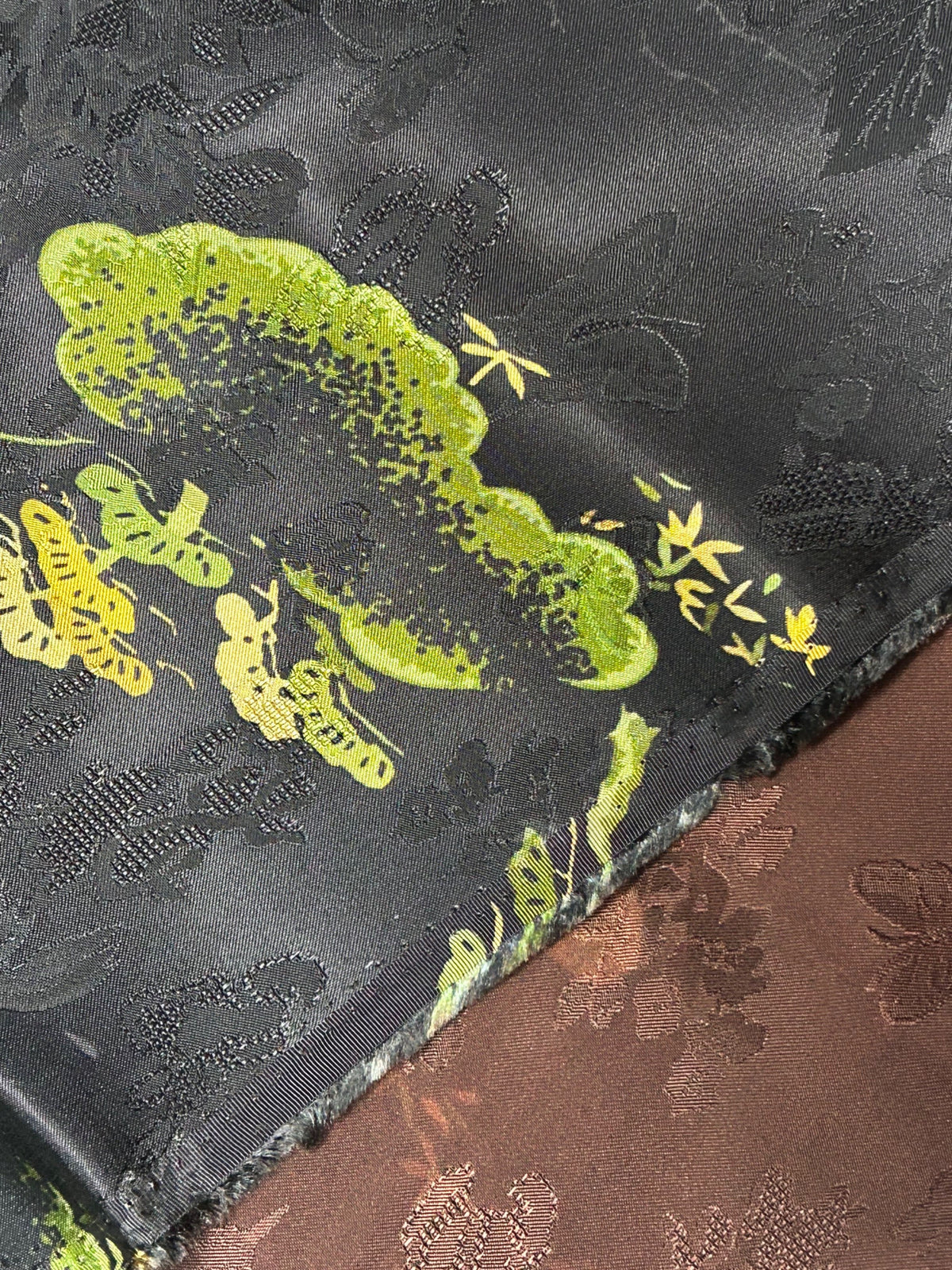 Close-up of overlapping dark charcoal and reddish-brown fabric swatches with raised floral patterns; the charcoal cloth has a bright lime-green and yellow botanical motif.