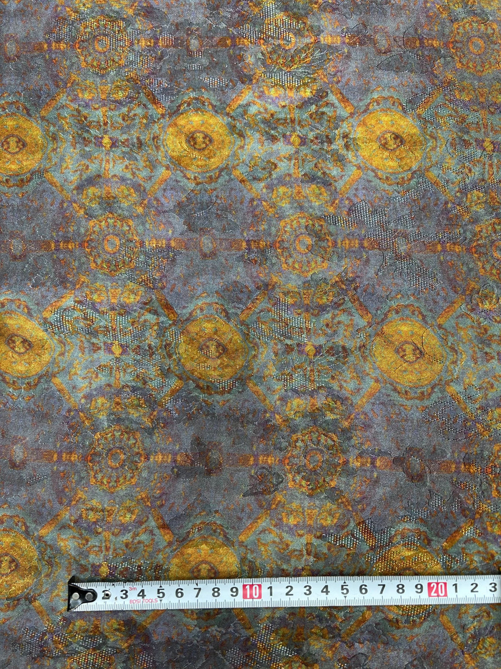 Close-up of blue-gray fabric with a repeating gold and rust floral medallion pattern and fine dotted accents; a measuring tape lies across the bottom.