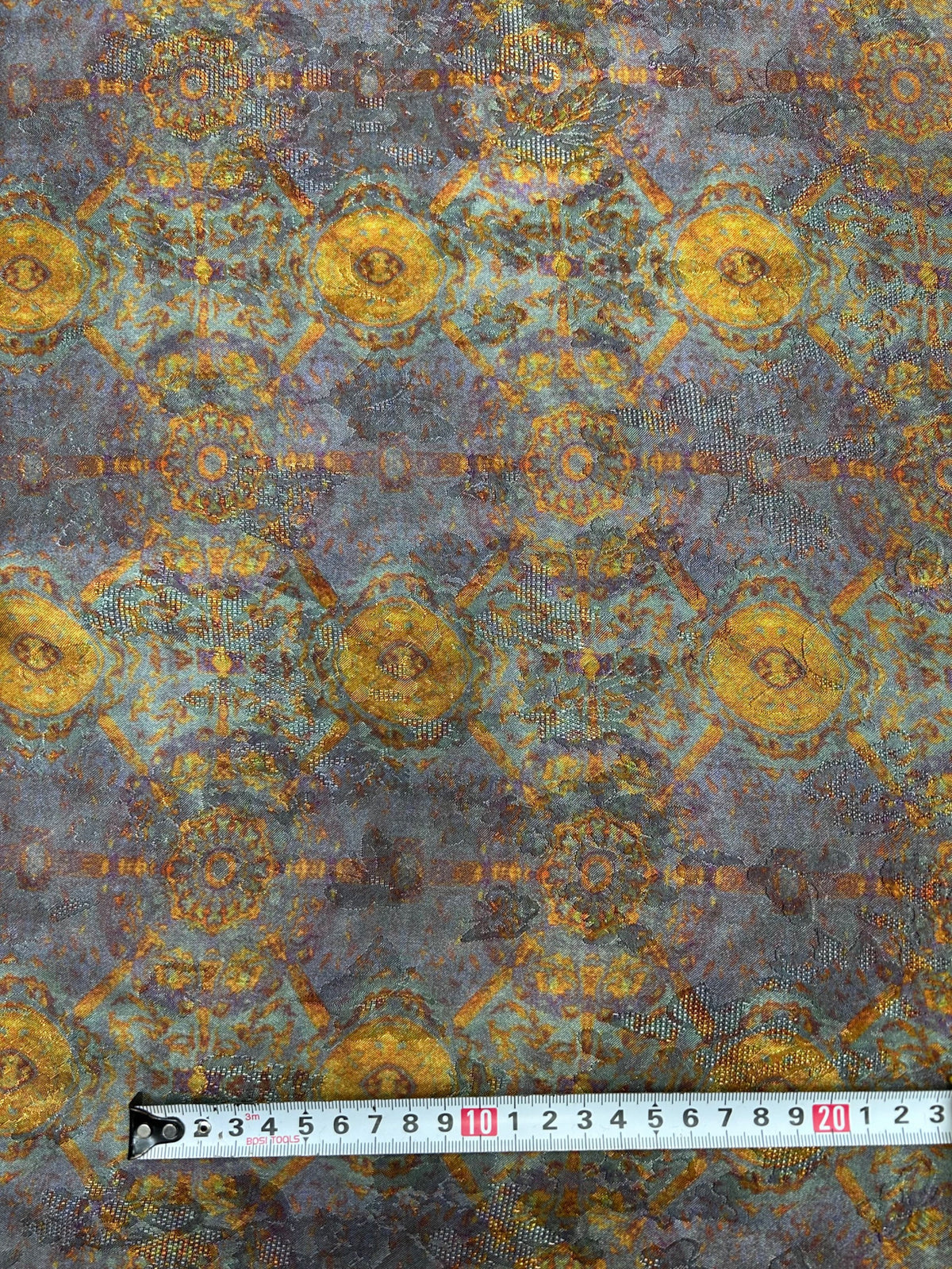 Close-up of blue-gray fabric with a repeating gold and rust floral medallion pattern and fine dotted accents; a measuring tape lies across the bottom.