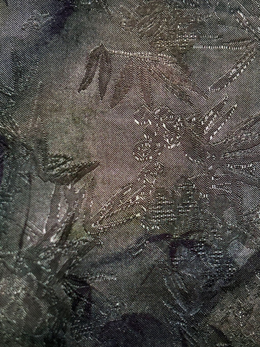 Close-up of dark charcoal cloth with a subtle woven pattern of overlapping leaves and large floral shapes, shown as a flat fabric surface.