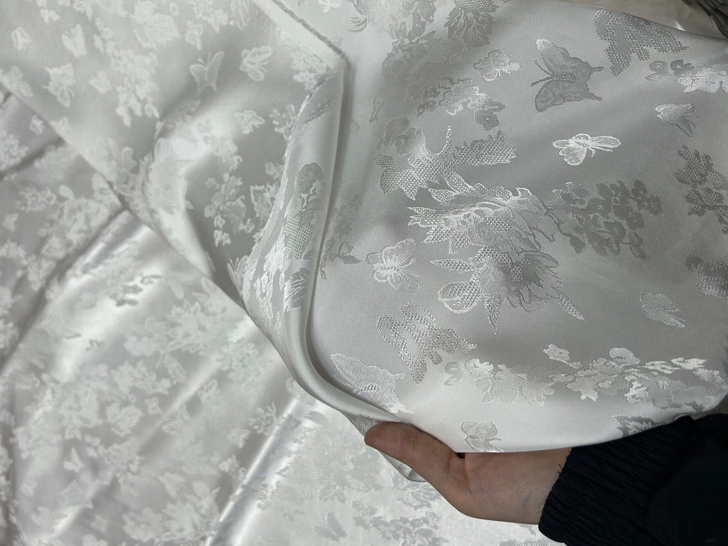 Close-up of glossy white cloth with a repeating floral and butterfly motif in white and gray; one section is folded back over the fabric.