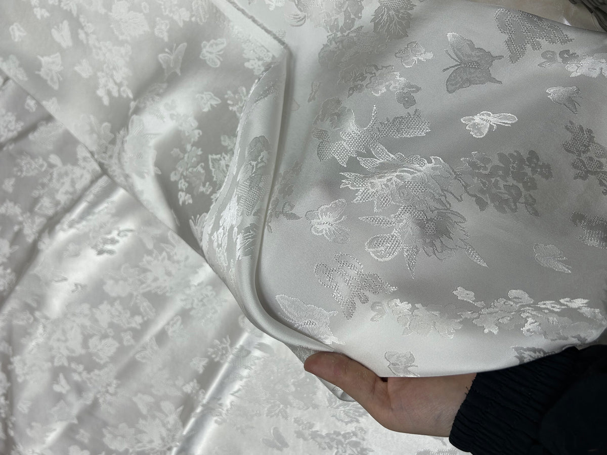 Close-up of glossy white cloth with a repeating floral and butterfly motif in white and gray; one section is folded back over the fabric.