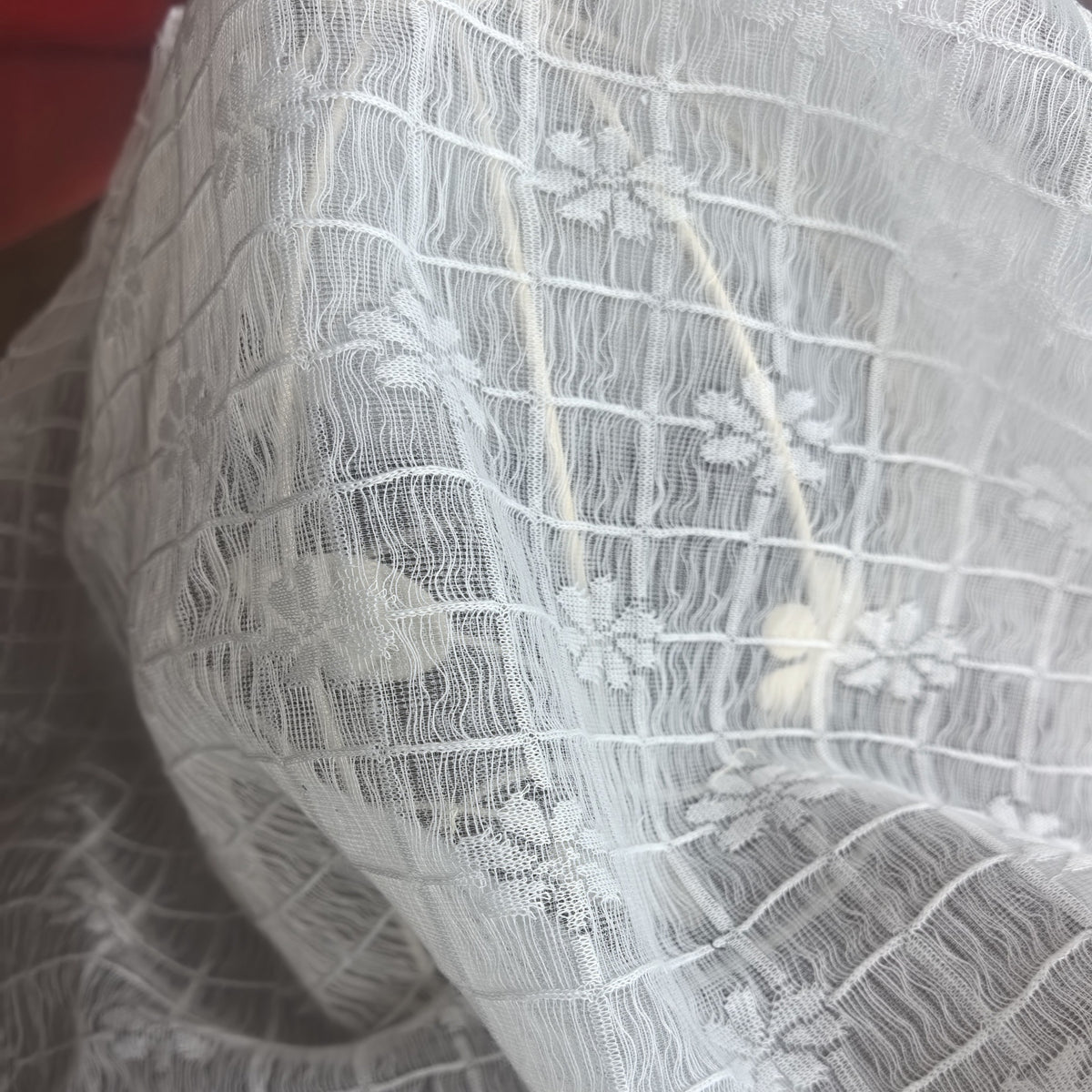 Close-up of white lace fabric with a repeating grid and small floral motifs, draped in curved folds.