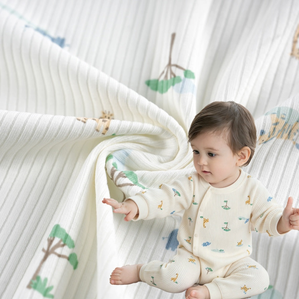 A baby wearing a cream ribbed outfit with small green, blue, and ochre motifs sits beside matching cloth draped in curved folds; close-up garment view.