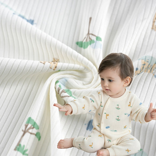 A baby wearing a cream ribbed outfit with small green, blue, and ochre motifs sits beside matching cloth draped in curved folds; close-up garment view.