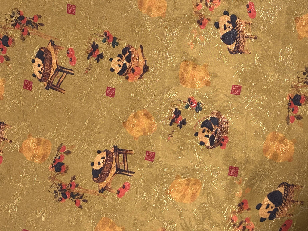 Close-up of gold cloth patterned with scattered stylized pandas, orange blossoms, dark branches, and small red square seals.