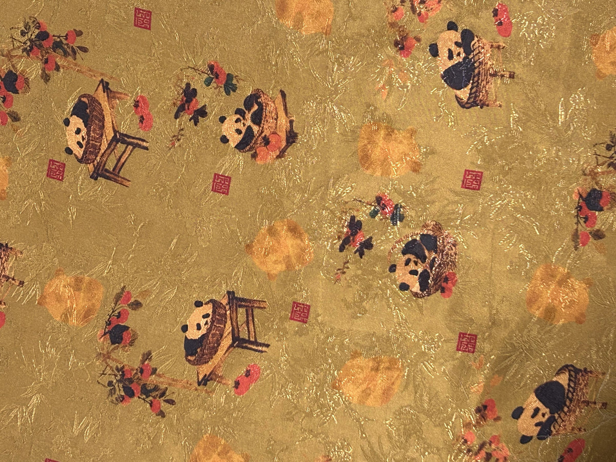 Close-up of gold cloth patterned with scattered stylized pandas, orange blossoms, dark branches, and small red square seals.