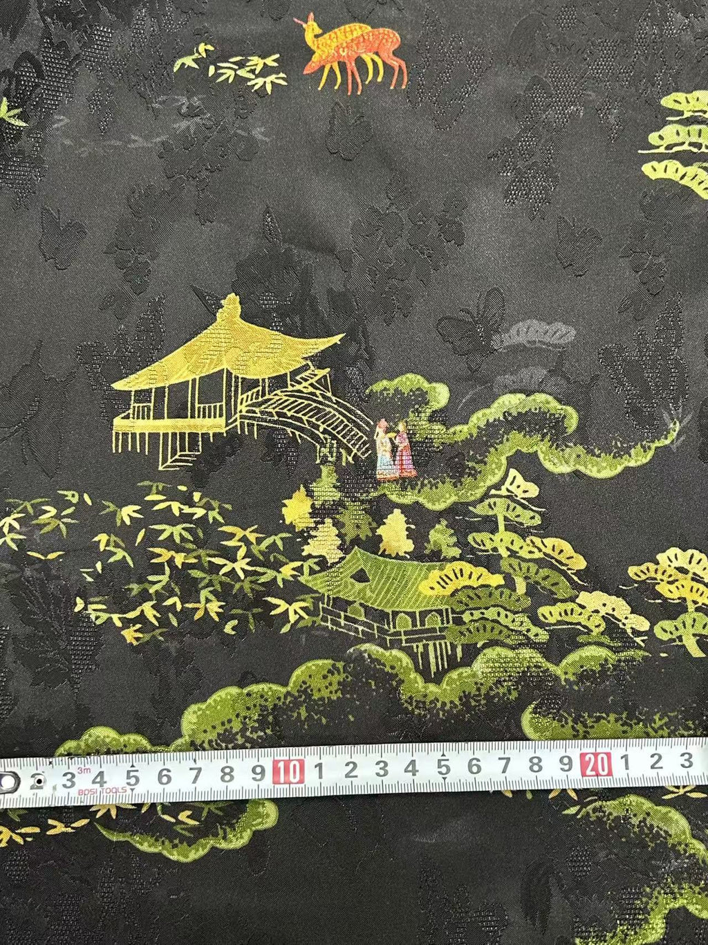 Close-up of black fabric printed with a textured floral pattern and colorful scenes of yellow pavilion buildings, green trees, small figures, and an orange deer. A measuring tape lies across the lower edge.