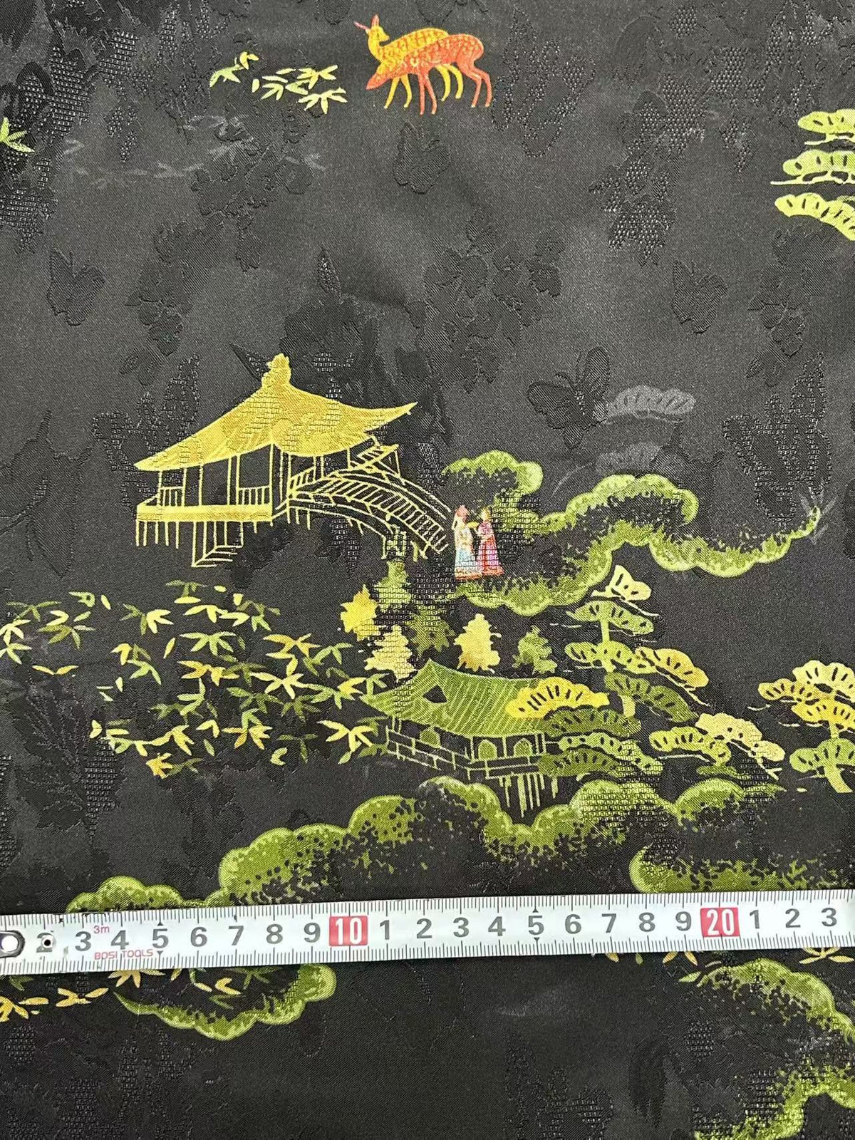 Close-up of black fabric printed with a textured floral pattern and colorful scenes of yellow pavilion buildings, green trees, small figures, and an orange deer. A measuring tape lies across the lower edge.