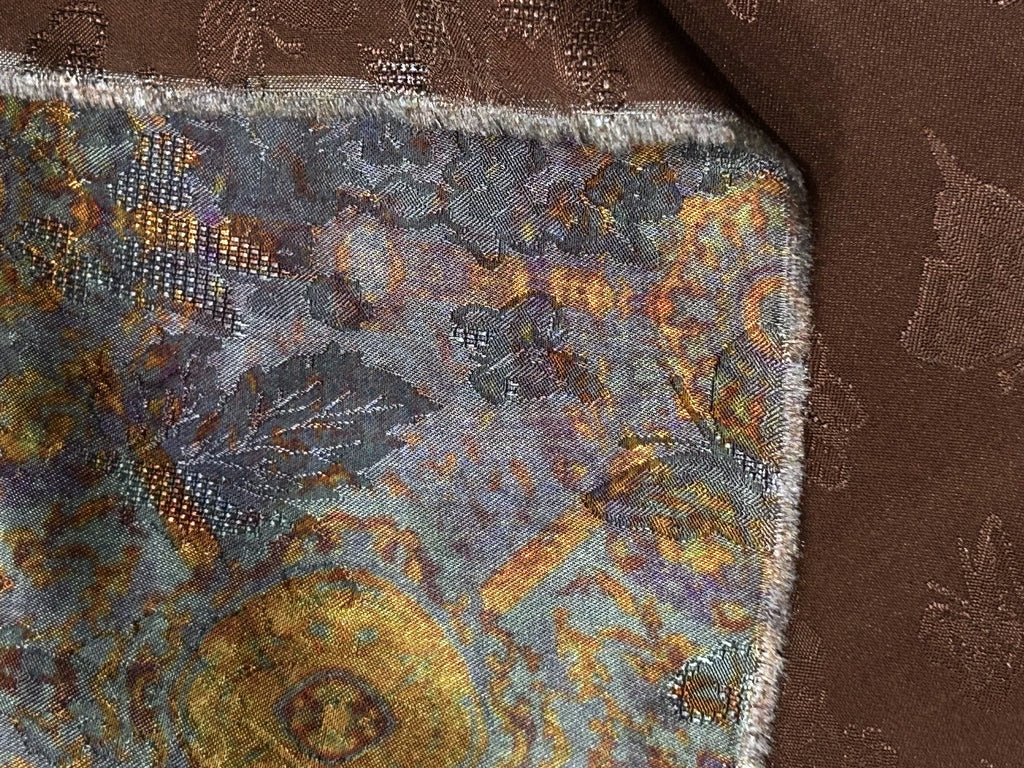 Close-up of a multicolored woven fabric swatch with a blue-gray, gold, and rust abstract motif, overlapped by a brown patterned cloth.