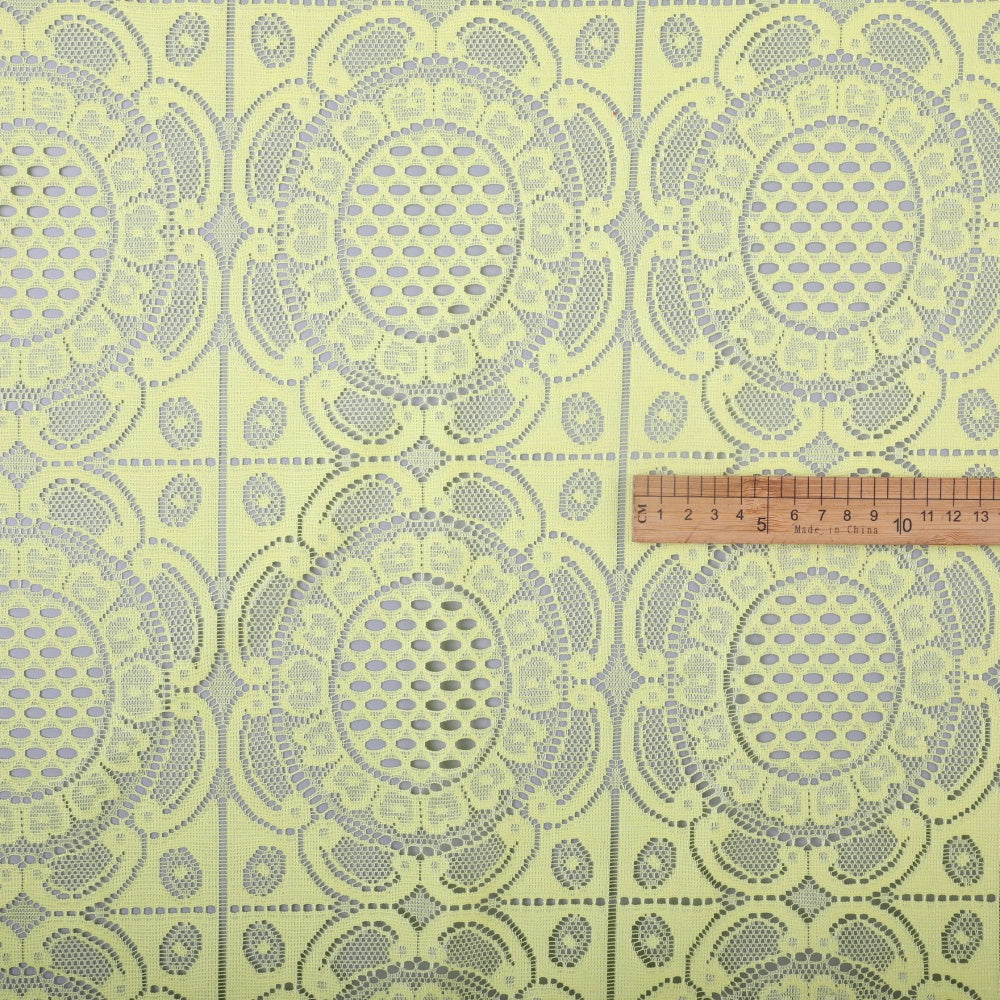 Pale yellow lace fabric lies flat, patterned with repeating large oval floral medallions, smaller blossoms, and openwork details. A ruler rests across the fabric near the right side.