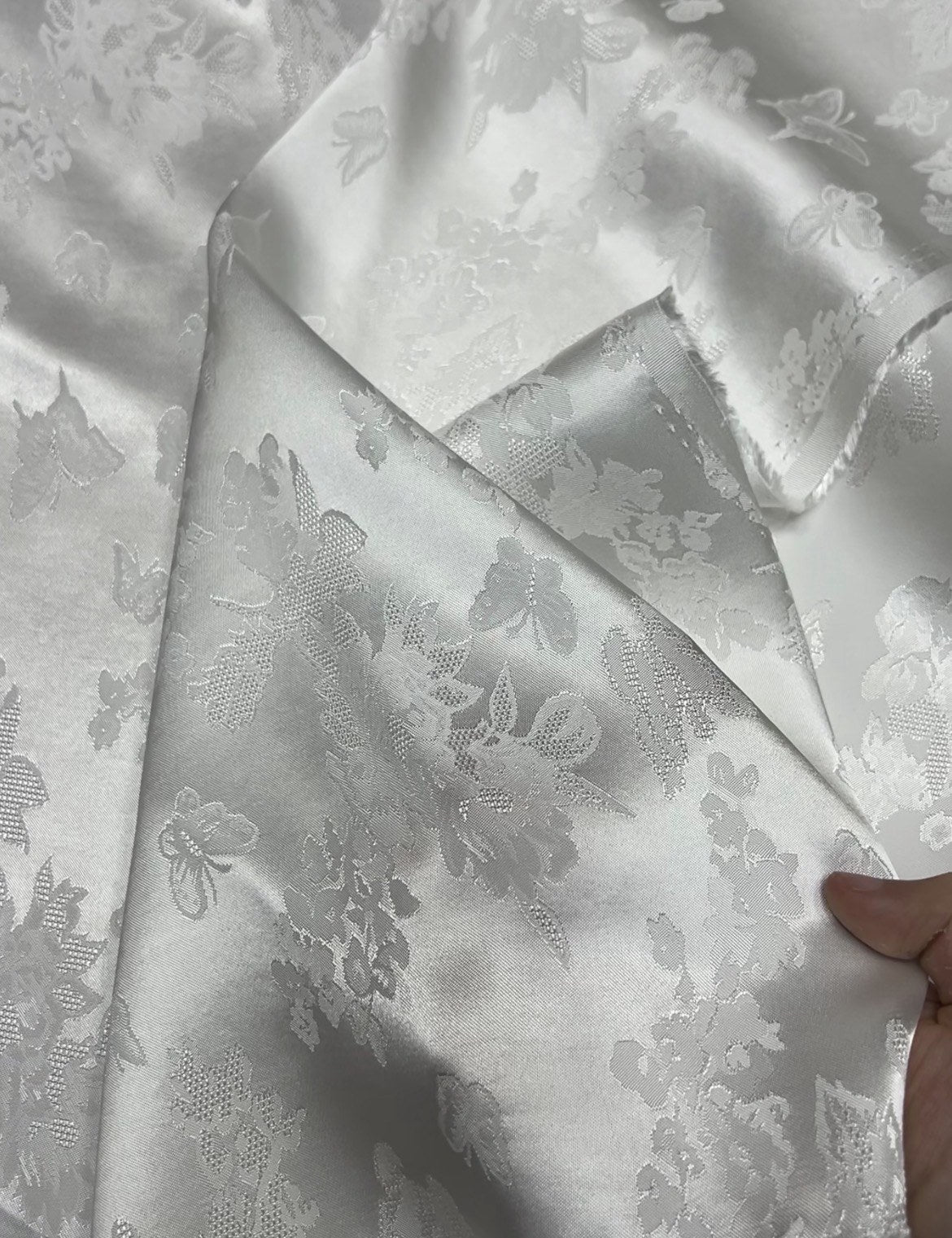 Close-up of folded ivory cloth with a subtle silvery floral and butterfly jacquard motif; a hand holds one corner.