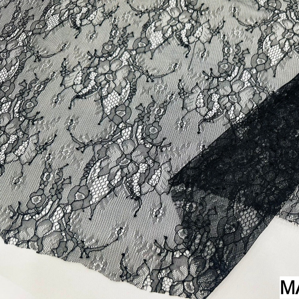 Black lace fabric lies flat, showing an intricate floral pattern with open mesh and scalloped edges against a light background.