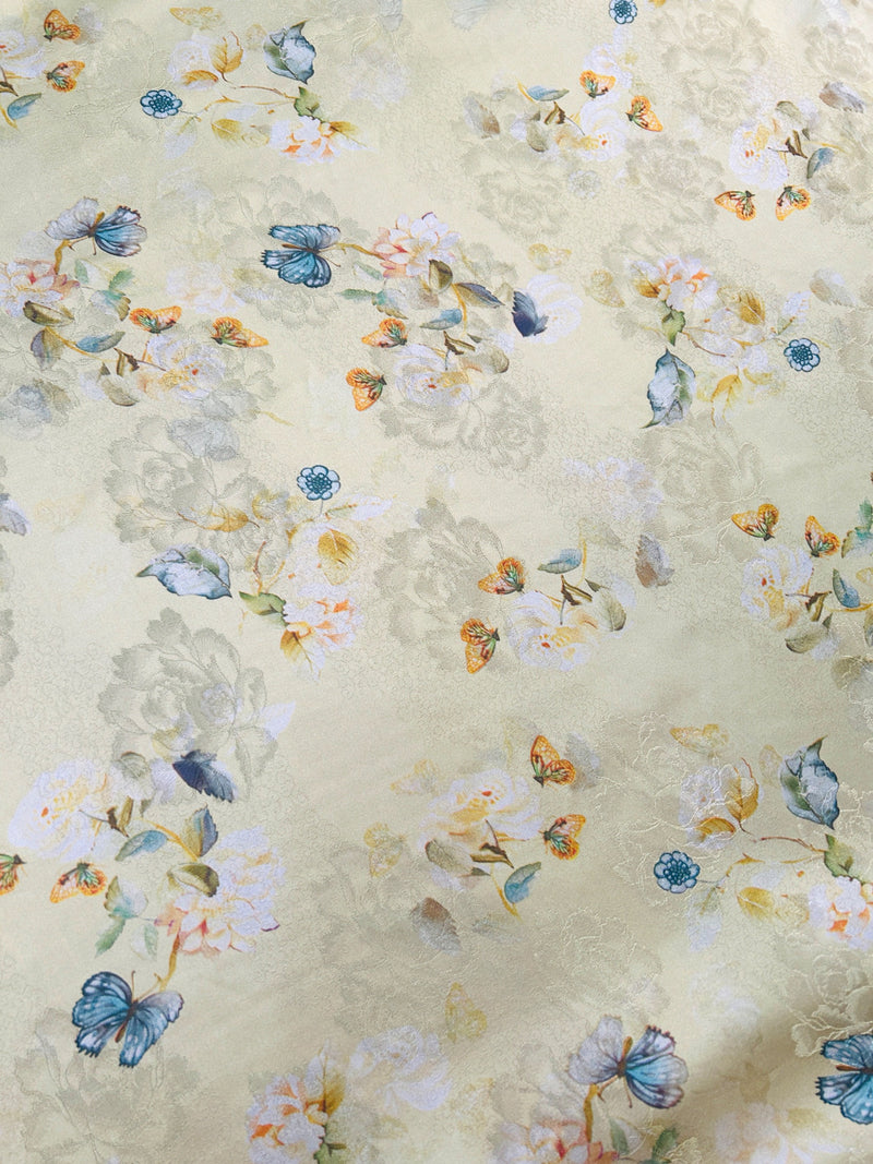 Close-up of pale cream fabric with a scattered floral pattern, blue butterflies, small teal flowers, and muted yellow, peach, and gray accents.