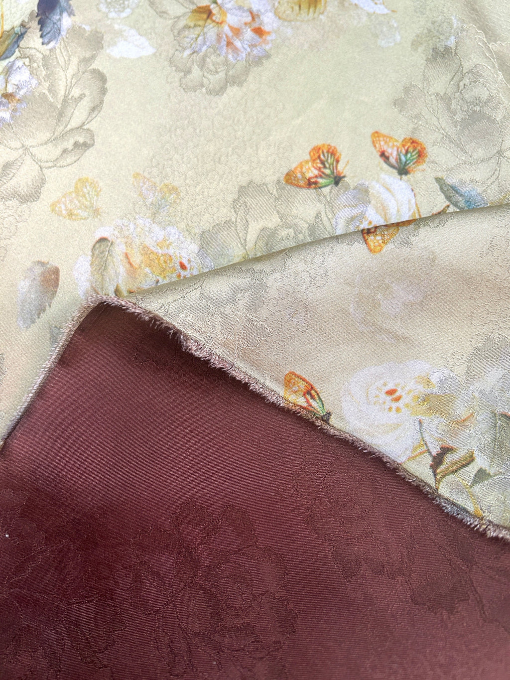 Close-up of folded fabric with a pale cream ground, muted floral outlines and orange, yellow, and blue butterfly motifs; a burgundy layer with a subtle floral pattern lies beneath it.