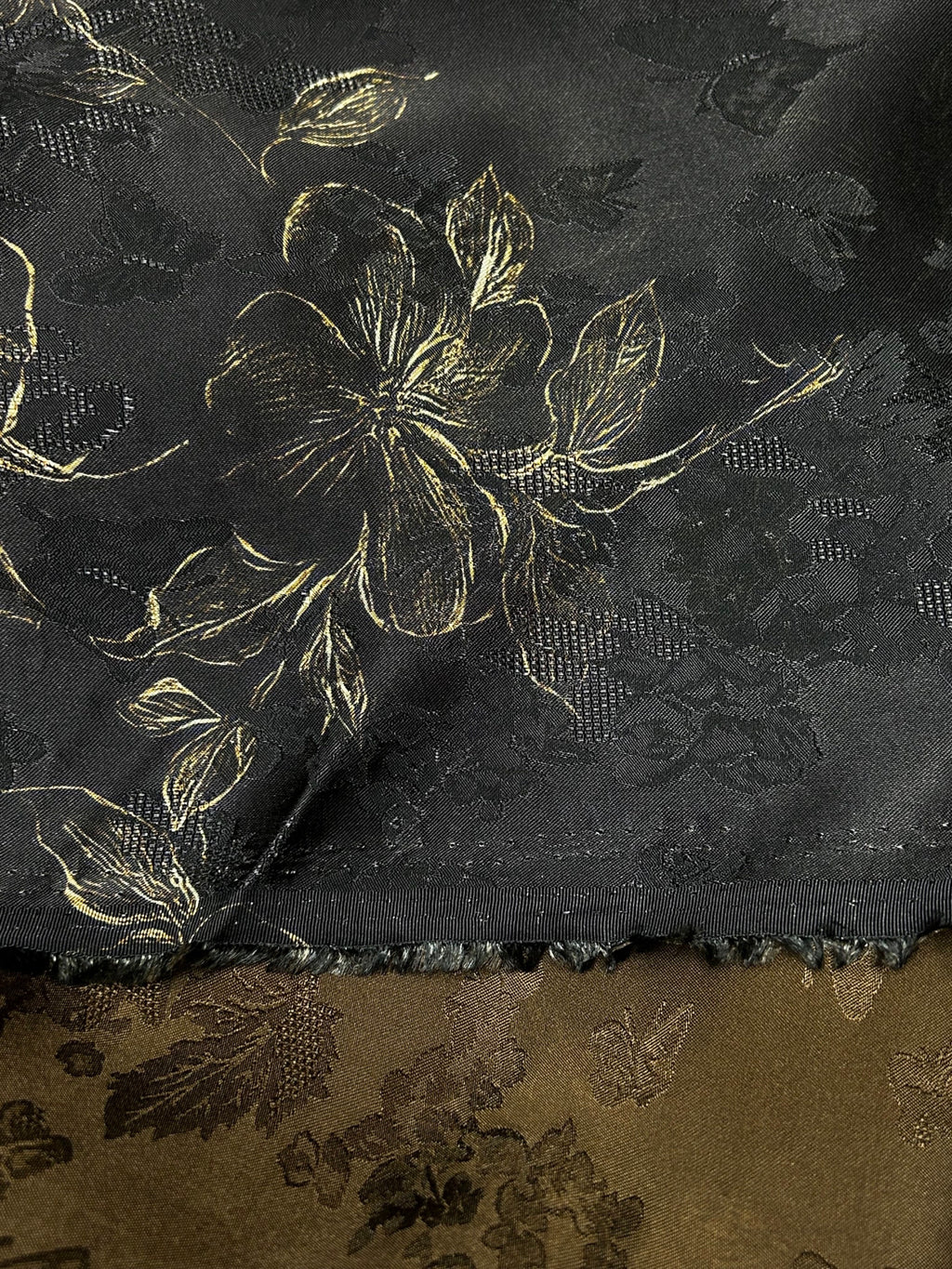 Close-up of two patterned fabric swatches: a black upper cloth with gold floral outlines and subtle dark woven motifs, above an olive-brown cloth with darker floral shapes; their edges meet across the image.