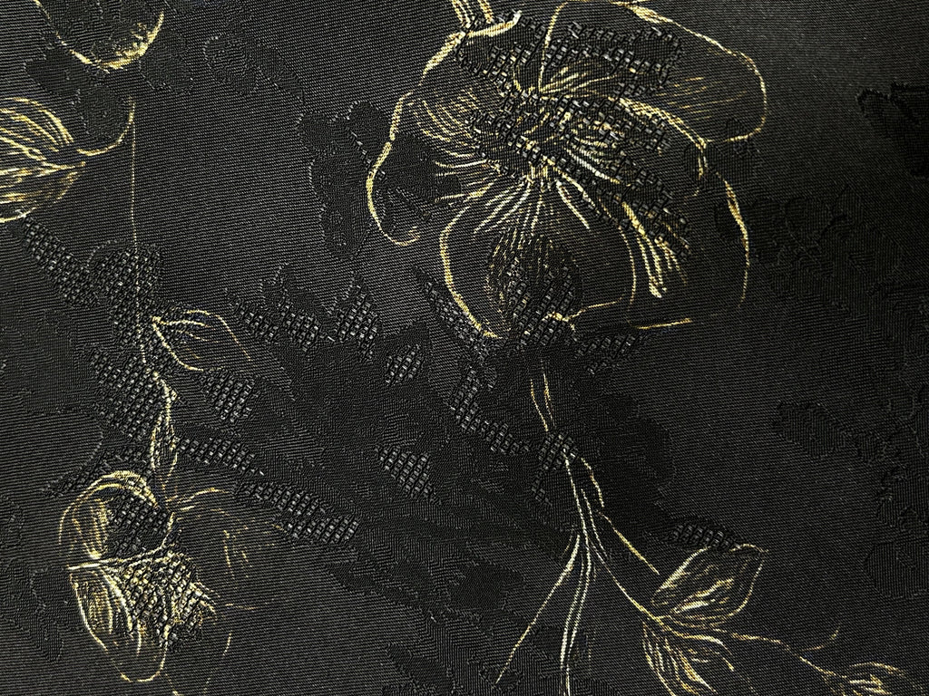 Close-up of black fabric with a dense woven floral motif in black and muted gold, showing large blossoms, leaves, and curling stems.