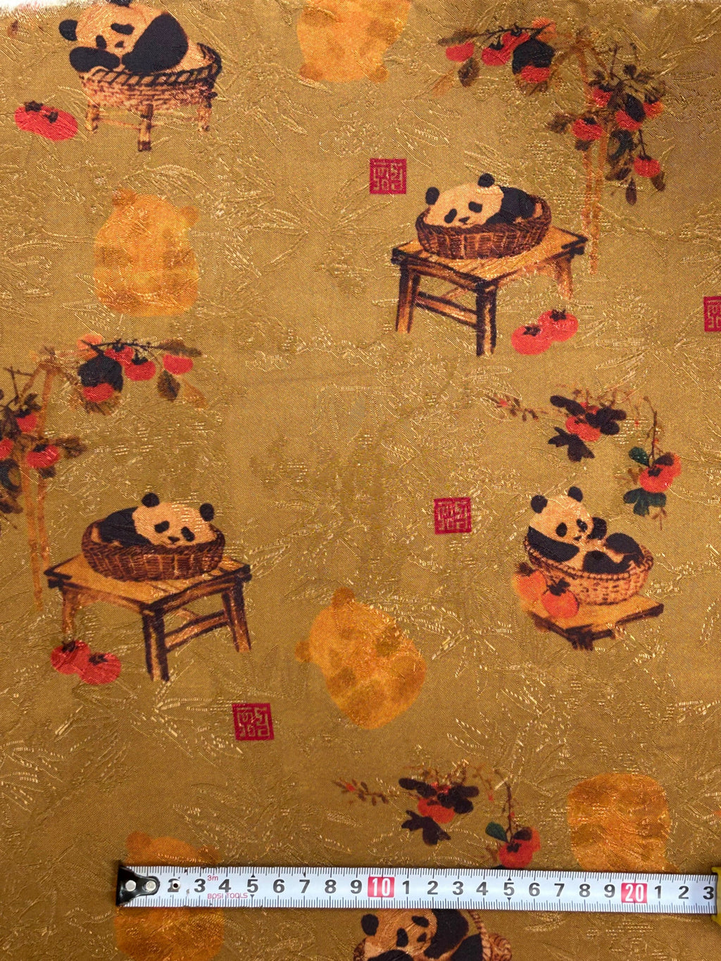 Close-up of gold cloth printed with pandas in woven baskets, some on small wooden tables, scattered among red and dark floral branches and red seal-like squares; a measuring tape runs along the bottom edge.