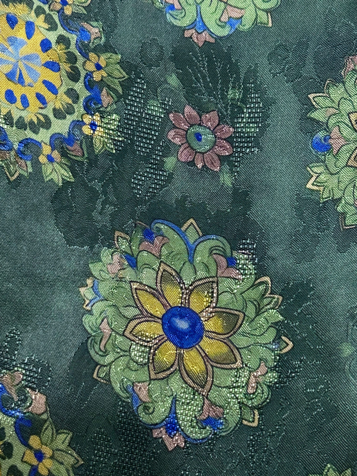 Close-up of dark green cloth with a repeating floral motif: large yellow-petaled flowers with bright blue centers among pale green leaves, smaller pink flowers, and blue accents.