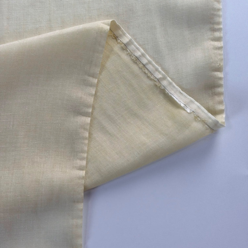 Close-up of pale ivory cloth folded back to show a triangular underside and a narrow stitched edge; the plain fabric has a fine grid texture and muted cream shading.