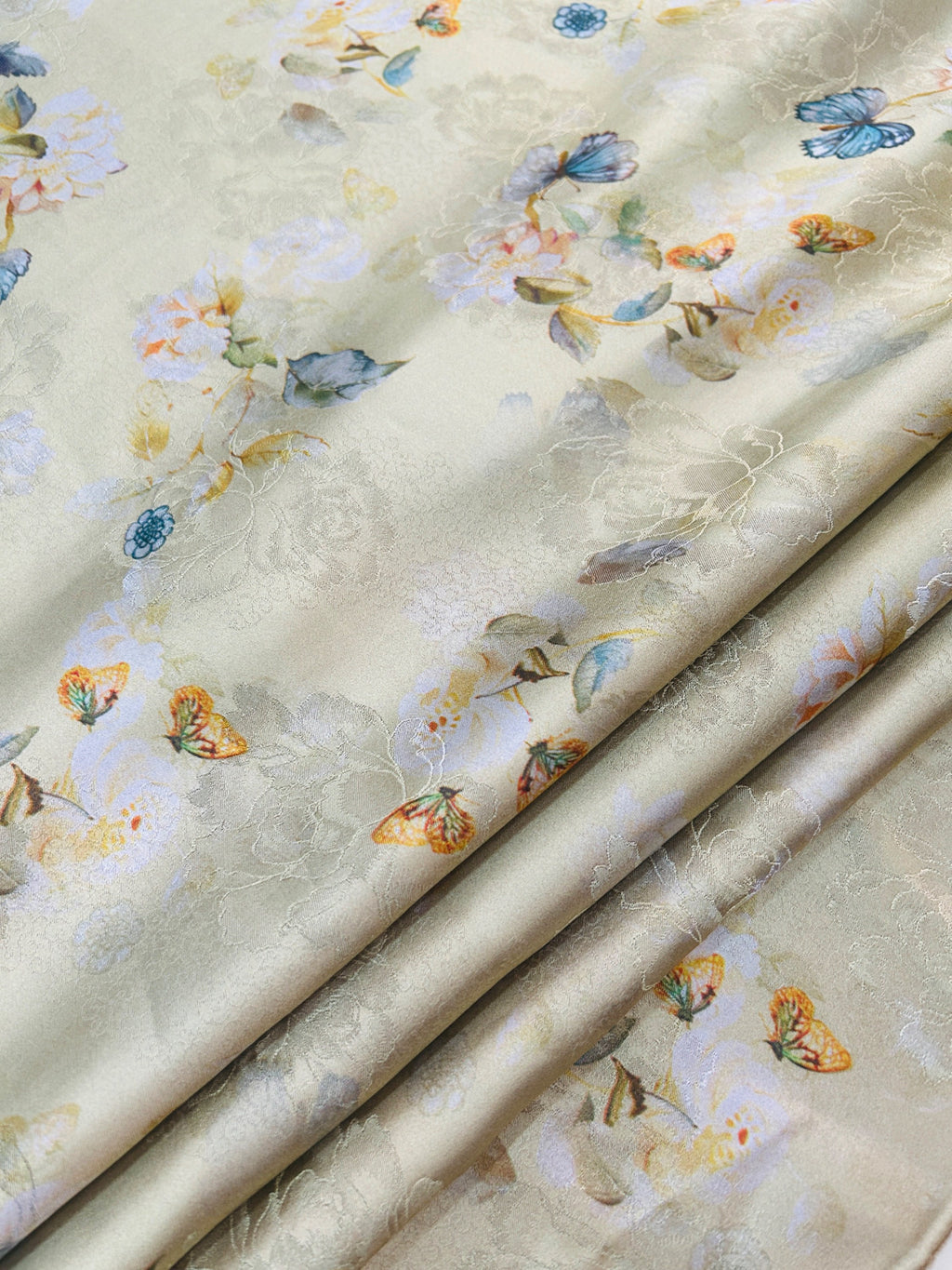 Close-up of folded cream fabric with a subtle woven floral pattern and scattered pastel flowers, leaves, and blue, orange, and yellow butterflies.