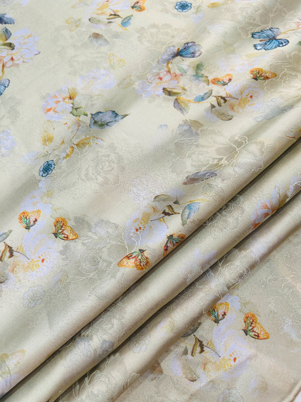 Close-up of folded cream fabric with a subtle woven floral pattern and scattered pastel flowers, leaves, and blue, orange, and yellow butterflies.