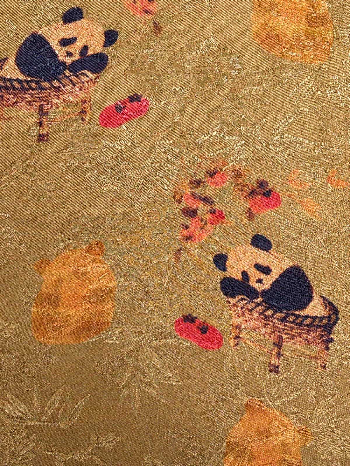 Close-up of gold-brown fabric with a textured woven pattern, scattered red and orange flowers, and two cartoon pandas seated in baskets; the cloth is shown flat.