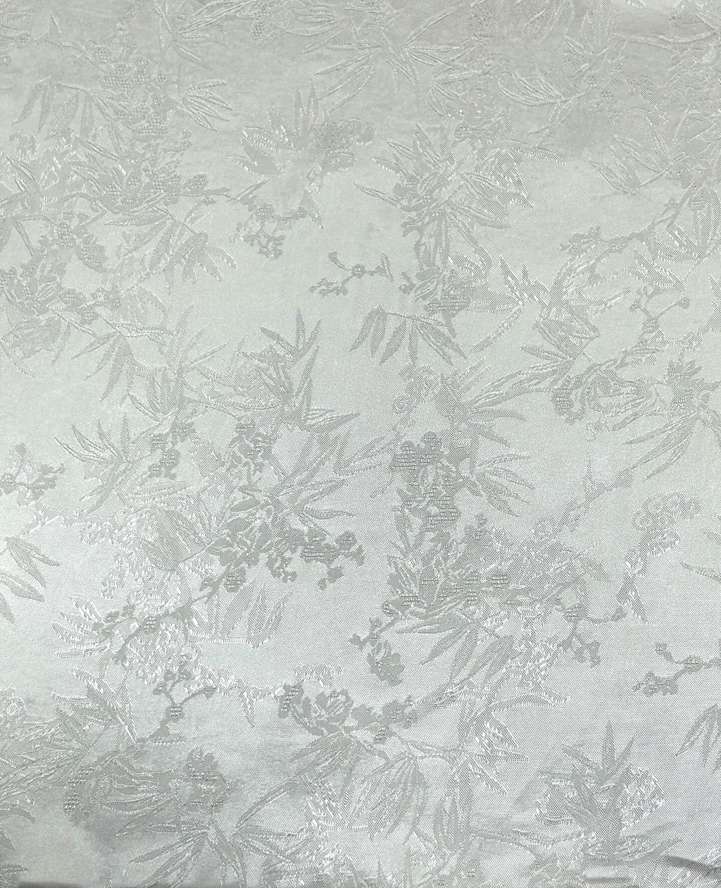Close-up of a pale gray fabric swatch with a repeating, scattered botanical motif of leafy branches and small blossoms woven in slightly darker gray and silvery tones.
