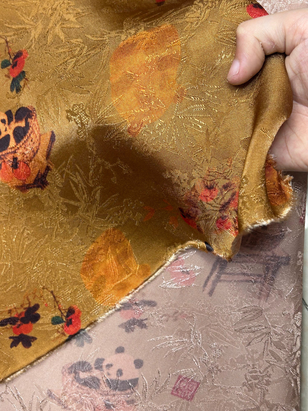 Close-up of a folded gold cloth with an ornate woven botanical pattern and scattered red, orange, and dark blue motifs; a hand holds up one edge.