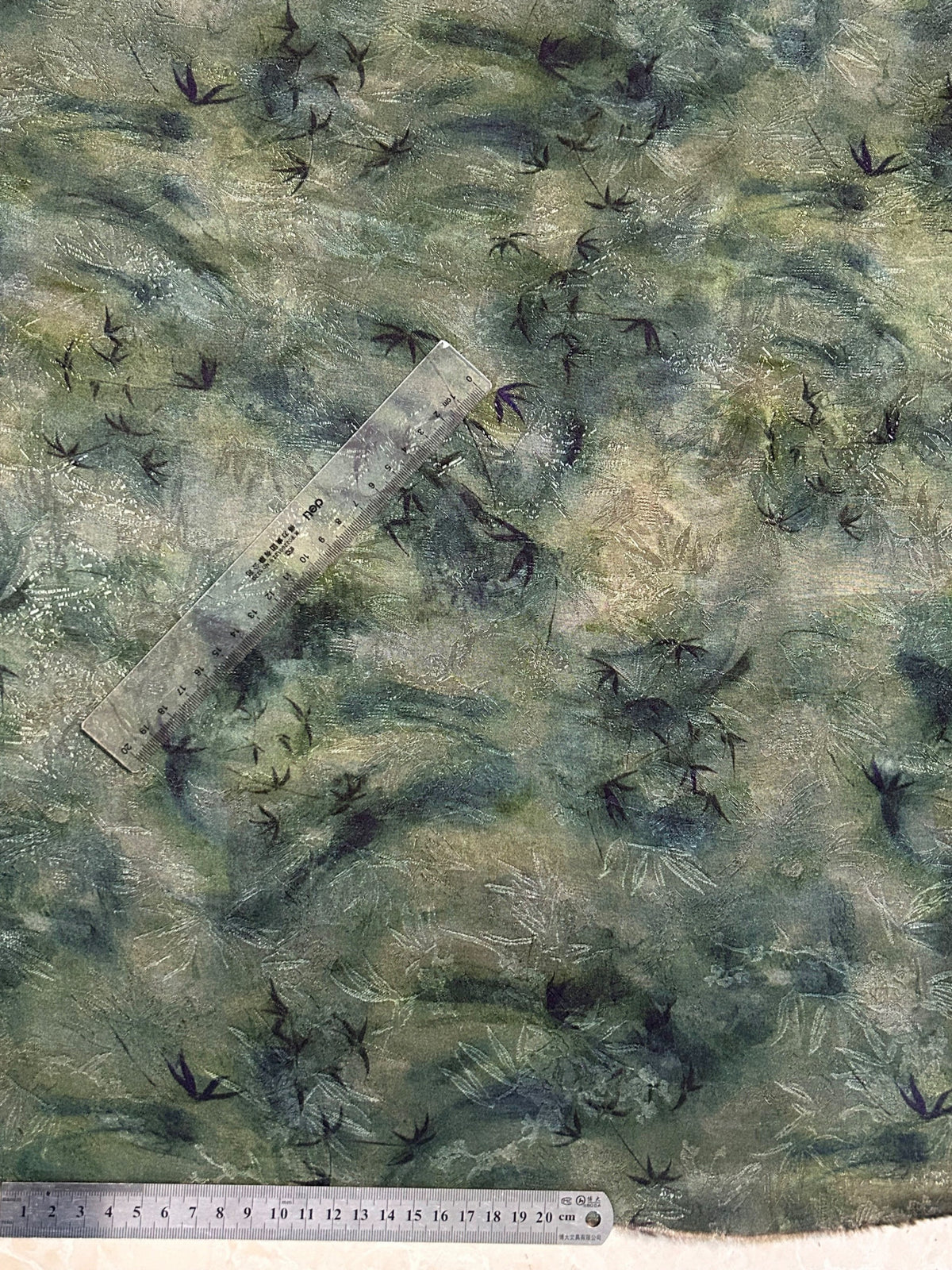 Close-up of a muted sage, gray, and beige fabric swatch with scattered dark bird silhouettes and fine pale leaf-like marks; two clear rulers lie across it.