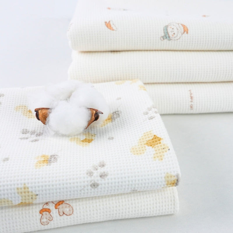 Cream white waffle-textured fabric with small pale yellow, gray, and orange motifs is folded in a layered stack. A loose tuft of white filling and a brown seed pod rest on the top printed piece.