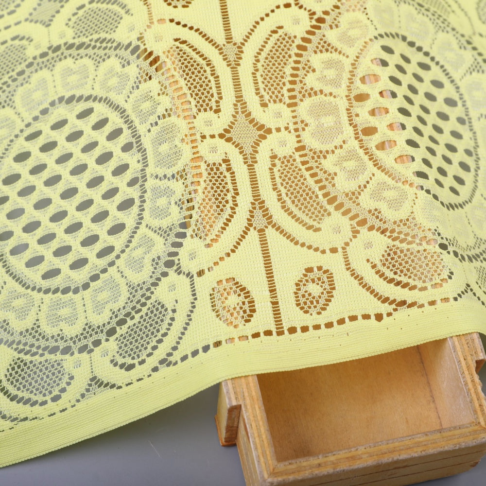 Pale yellow lace fabric with a repeating openwork floral and geometric motif, shown in a close view draped over a wooden tray.