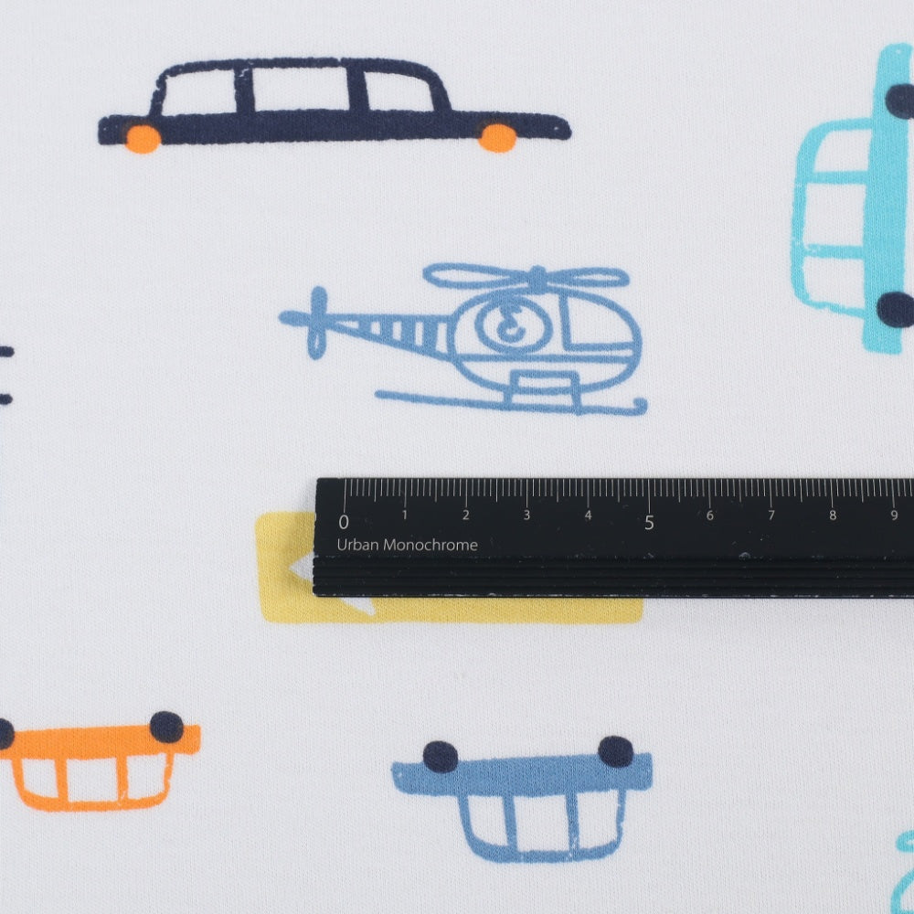 Close-up of white fabric printed with colorful vehicle motifs, including a navy limousine, blue helicopter, and partial cars; a black ruler lies across the center.