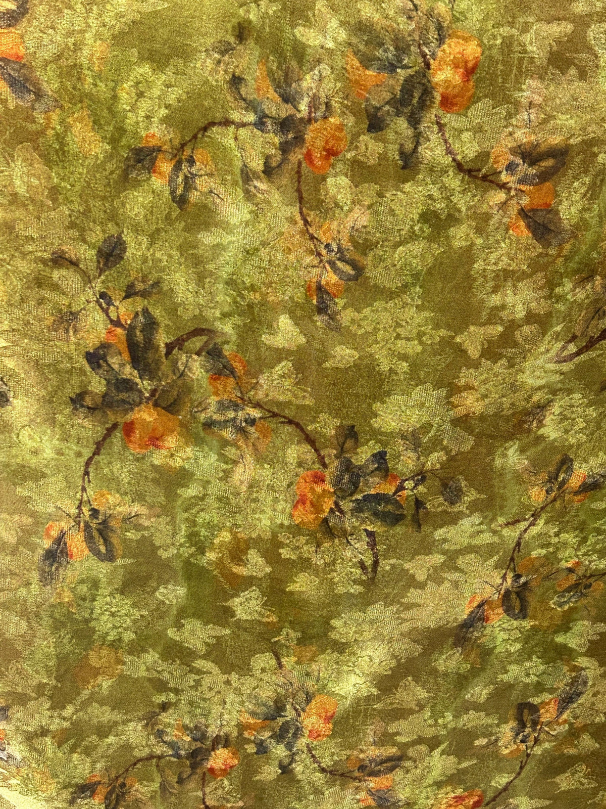 Close-up of olive green fabric with a dense, irregular floral motif of muted yellow-green leaves, dark brown branches, and scattered rust-orange blossoms.