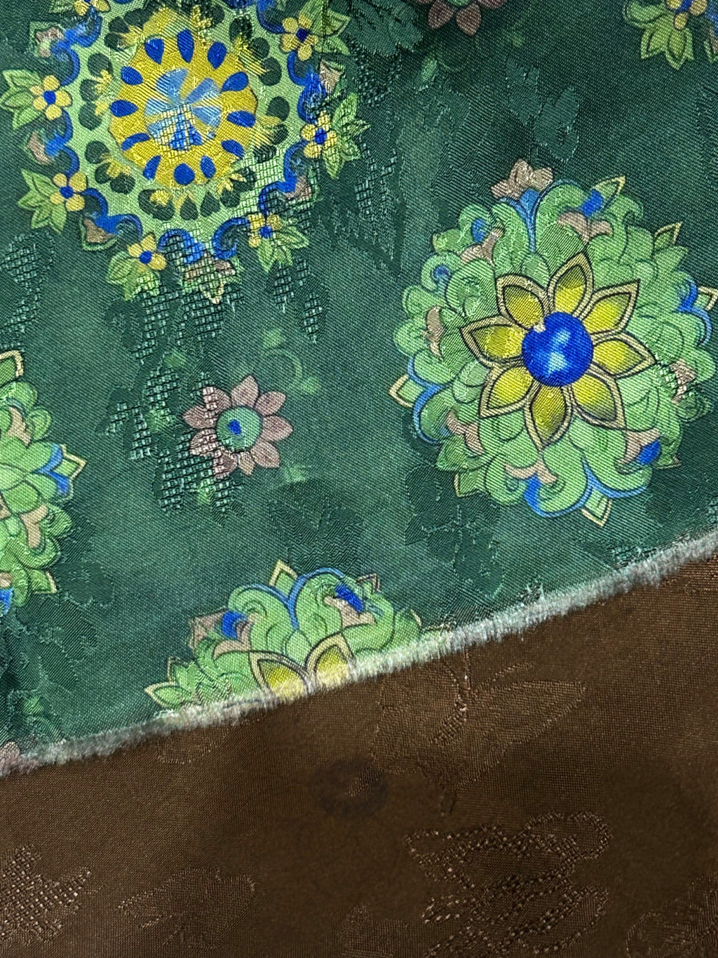 Close-up of a dark green cloth swatch with scattered floral motifs in lime green, yellow, blue, and pale pink; a fuzzy light edge separates it from brown fabric beneath.