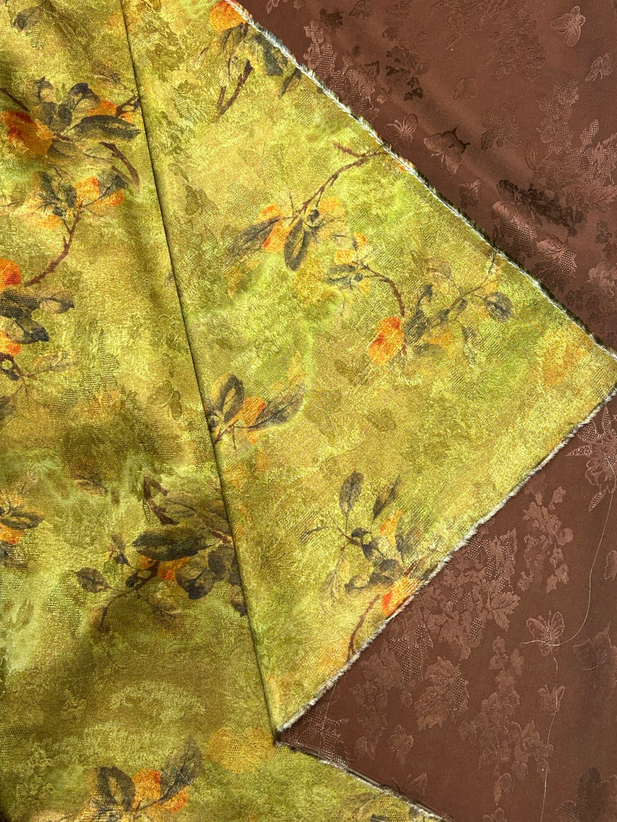 Close-up of folded cloth with a mottled yellow-green ground, scattered dark leafy branches and small orange accents; a brown floral-patterned fabric shows beneath it.