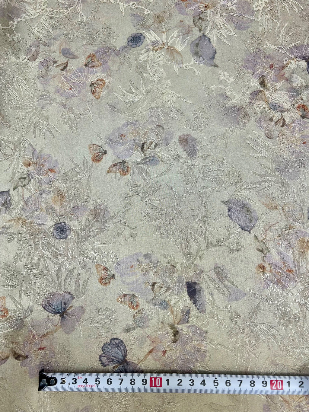 Close-up of pale gray-green fabric with a dense, tonal embroidered pattern of leaves and branches, scattered muted purple, blue, and rust floral motifs, and a measuring tape along the bottom edge.