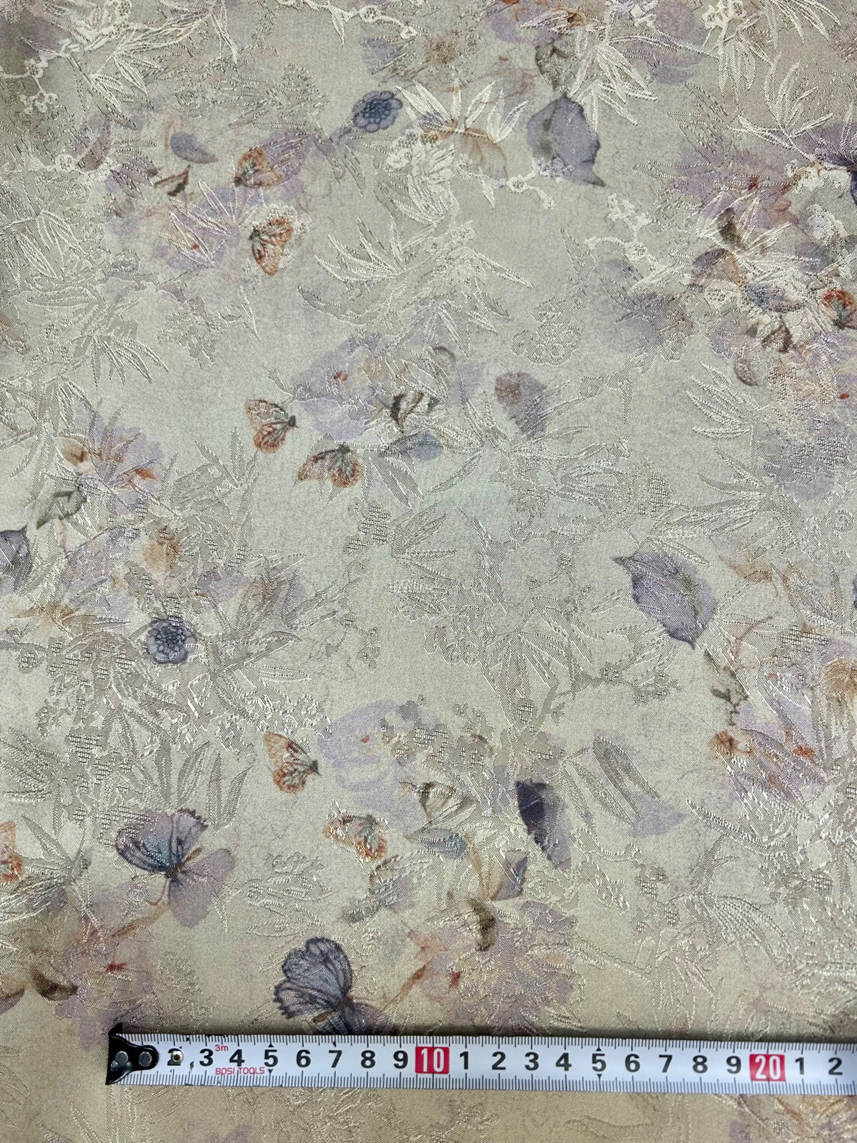 Close-up of pale gray-green fabric with a dense, tonal embroidered pattern of leaves and branches, scattered muted purple, blue, and rust floral motifs, and a measuring tape along the bottom edge.