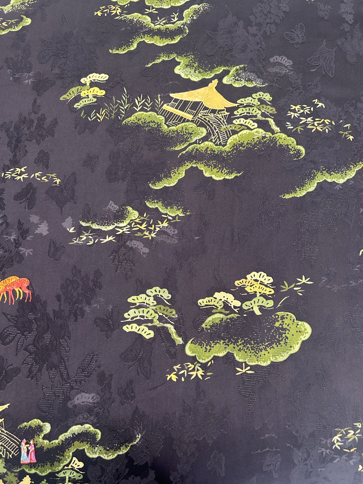 Close-up of dark charcoal fabric with a repeating pattern of yellow-green clouds, pine trees, bamboo sprigs, and small gold pagodas; a partial red-orange figure appears at the left edge.