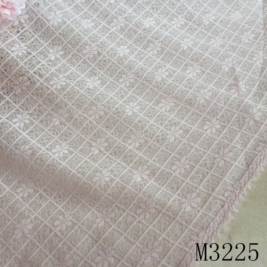 Close-up of pale blush lace fabric with a diagonal grid and repeating floral motifs; a pink flower is partly visible at the upper left, and the cloth edge runs diagonally across the lower right.