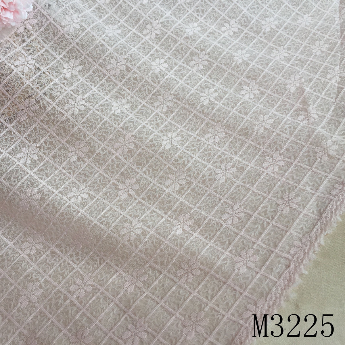 Close-up of pale blush lace fabric with a diagonal grid and repeating floral motifs; a pink flower is partly visible at the upper left, and the cloth edge runs diagonally across the lower right.